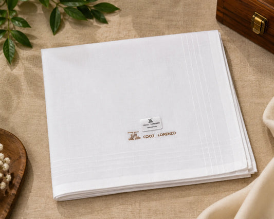 A folded white linen napkin with subtle grid lines and an embroidered logo reading COCCA LORENZO, styled on a beige fabric surface with decorative leaves and a wooden box.