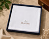 A folded white handkerchief with subtle grid lines and gold embroidered branding, presented inside a dark navy gift box on a beige fabric surface with decorative leaves and dried flowers.