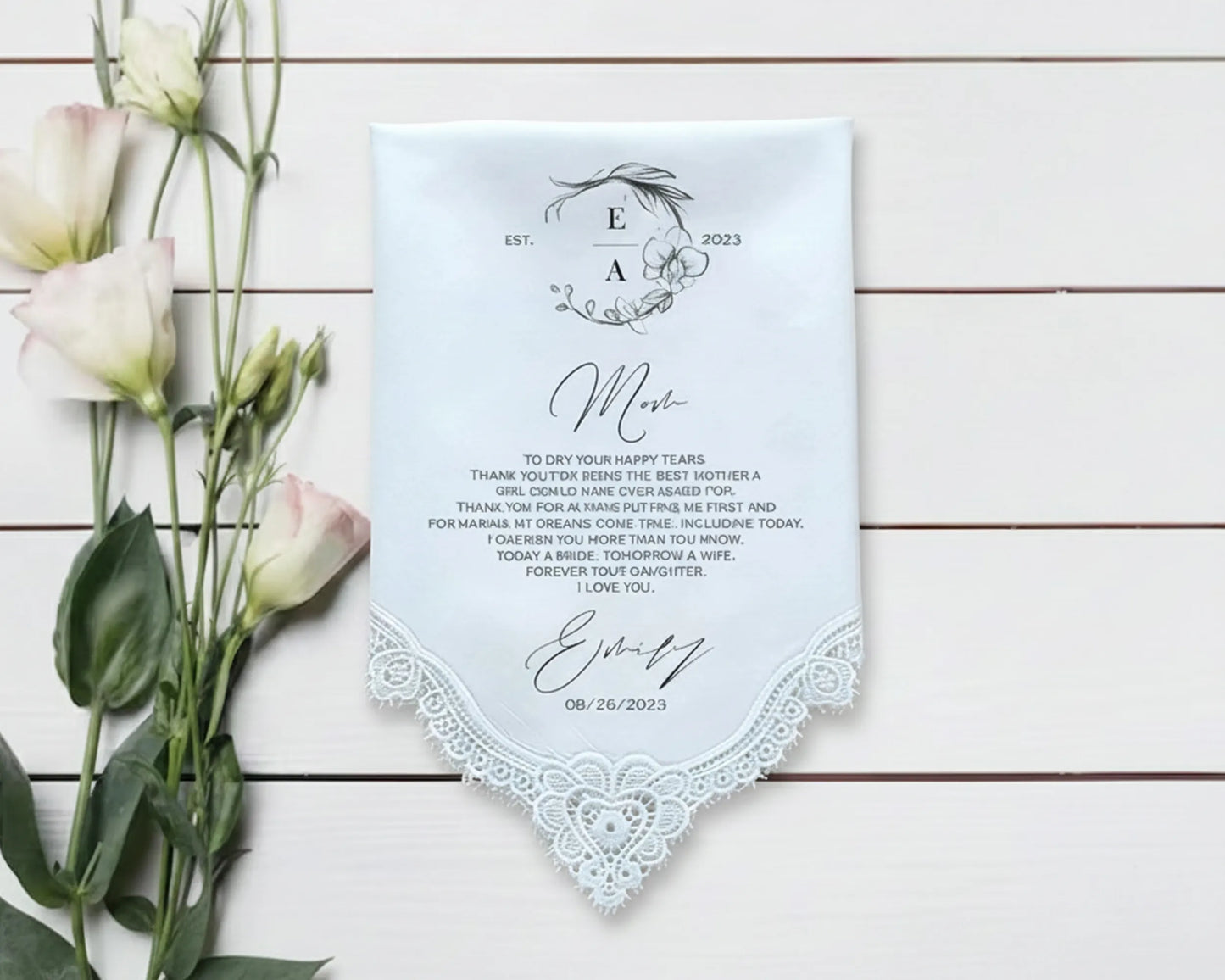 Light blue handkerchief with lace trim and personalized text on a wooden surface with flowers.