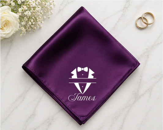 Purple pocket square with a tuxedo design and 'James' text on a marble surface with flowers and rings.