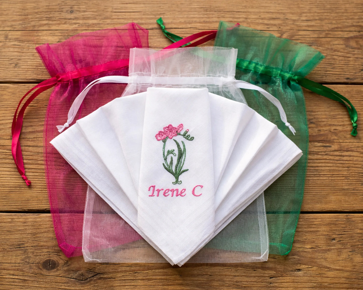 Set of embroidered handkerchiefs with floral design and text, surrounded by colorful drawstring bags on a wooden surface.