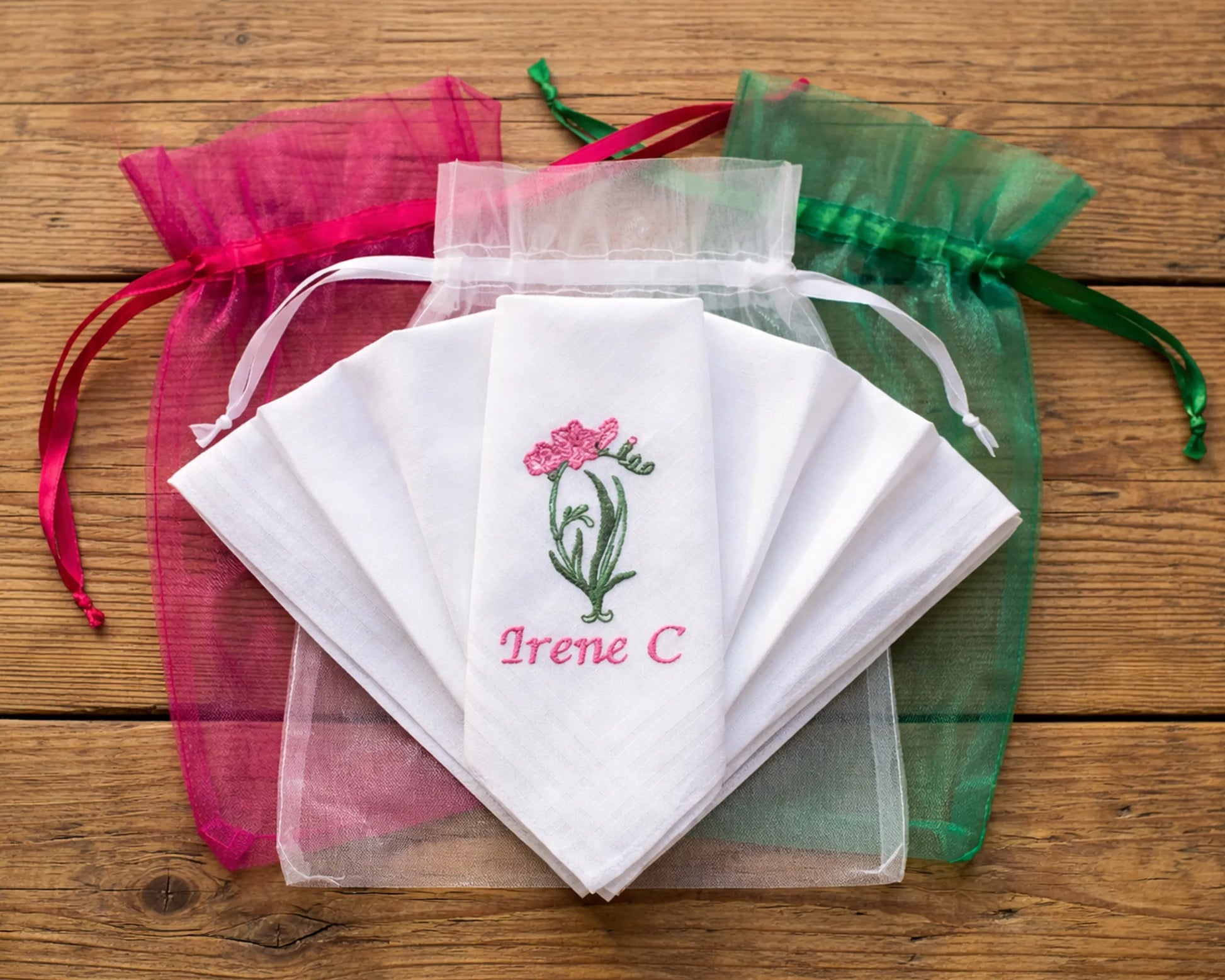 Set of embroidered handkerchiefs with floral design and text, surrounded by colorful drawstring bags on a wooden surface.