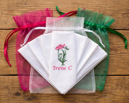 Set of embroidered handkerchiefs with floral design and text, surrounded by colorful drawstring bags on a wooden surface.