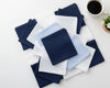 Set of folded blue, white, and gray hanky on a light surface with a coffee cup and plant.