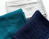 Set of folded blue, white, and gray hanky on a light surface with a coffee cup and plant.