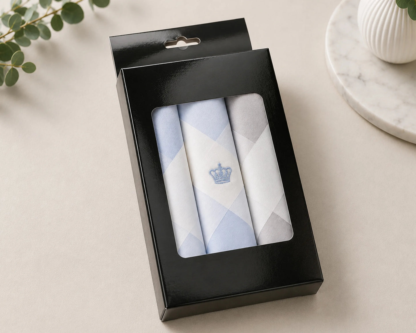 Set of three folded hanky in a black box on a light surface with a plant in the corner.
