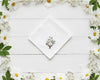 Centered embroidery of white daisies and green leaves on a white cotton handkerchief framed by real flowers