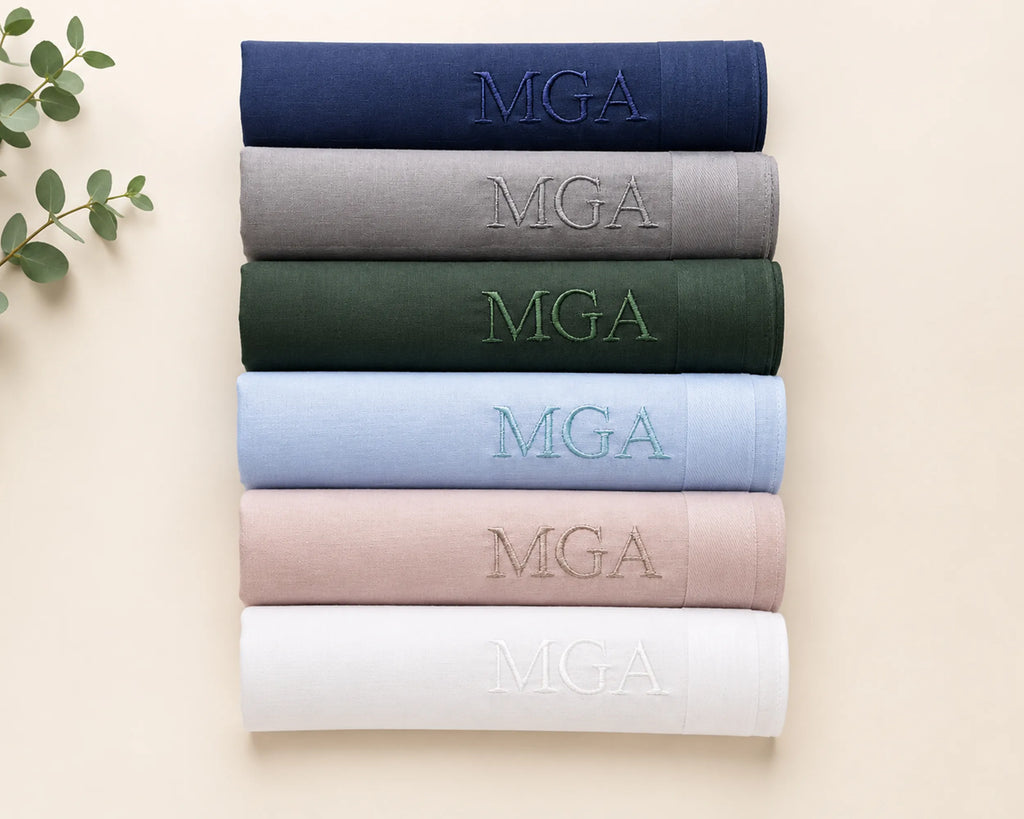 Stack of folded hanky with 'MGA' embroidery in various colors on a beige background.