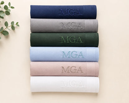 Stack of folded hanky with 'MGA' embroidery in various colors on a beige background.