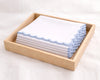 Stack of white handkerchiefs with blue scalloped edge in a wooden tray on a light background