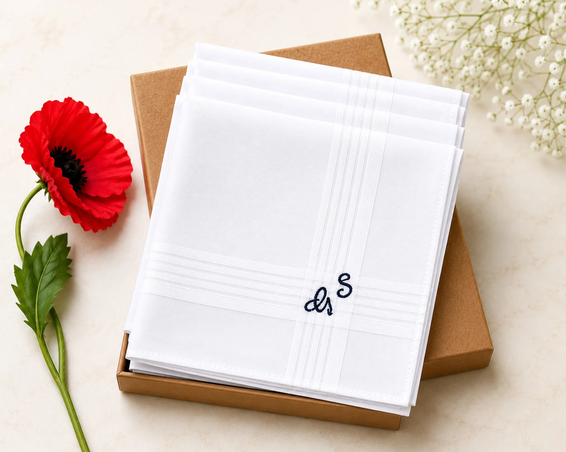Stack of white hanky with a monogram on a brown box, accompanied by a red flower and white flowers.