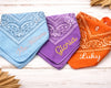Three bandanas in blue, purple, and orange with embroidered names on a wooden surface.
