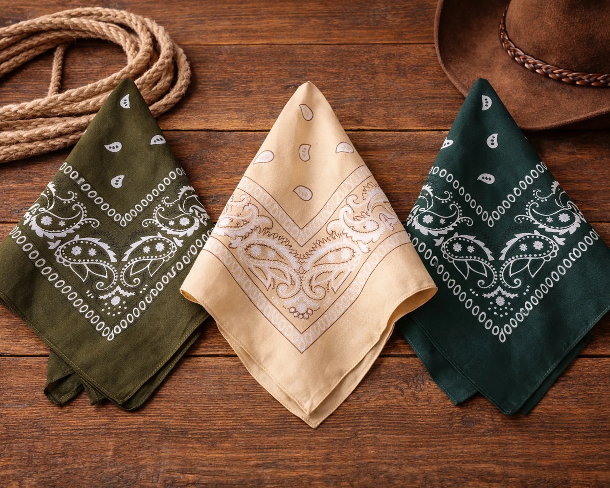 Three bandanas in green, beige, and dark green on a wooden surface with a cowboy hat and rope.