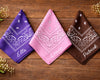 Three bandanas in purple, pink, and brown with names printed on them, placed on a wooden surface.