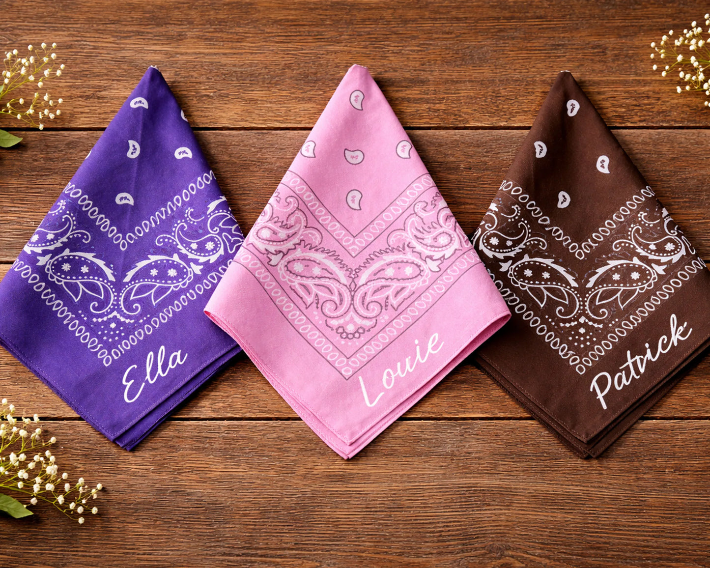 Three bandanas in purple, pink, and brown with names printed on them, placed on a wooden surface.