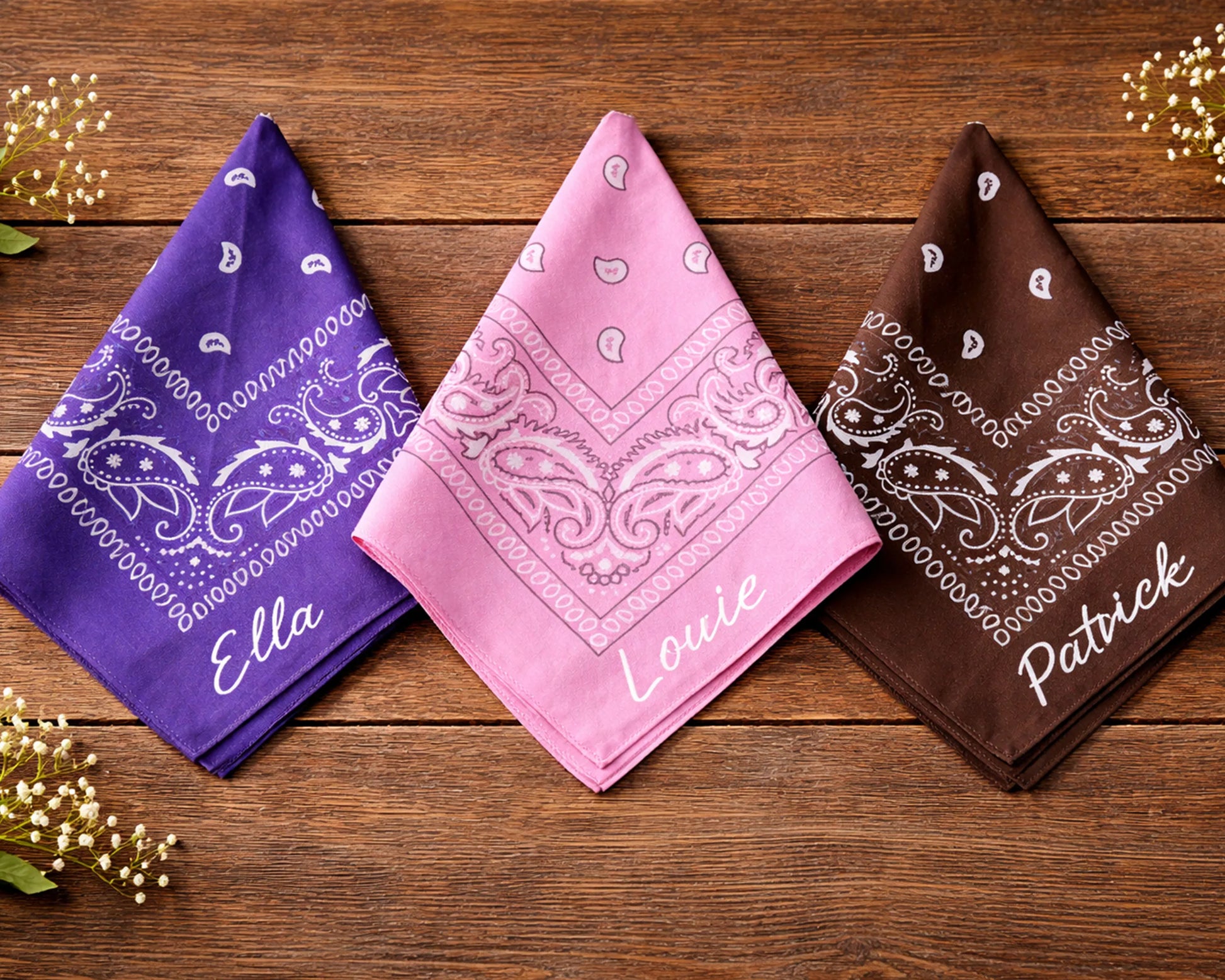 Three bandanas in purple, pink, and brown with names printed on them, placed on a wooden surface.