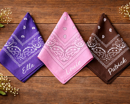 Three bandanas in purple, pink, and brown with names printed on them, placed on a wooden surface.
