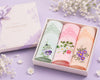 Three floral embroidered handkerchiefs in a box on a purple background with flowers.