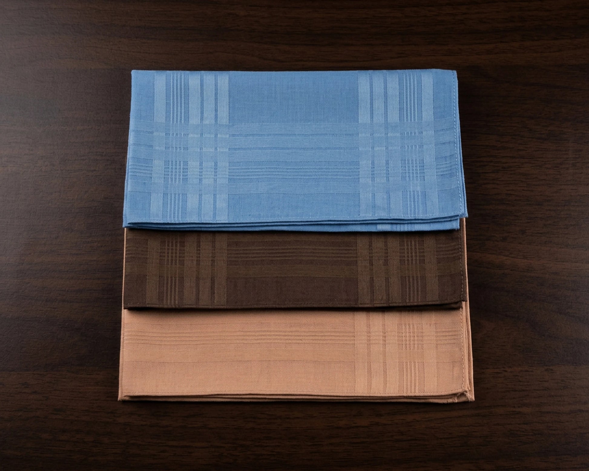 Three folded cotton hanky in blue, brown, and beige on a wooden surface
