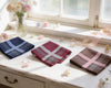 Three folded handkerchiefs on a white surface with flowers and a window in the background