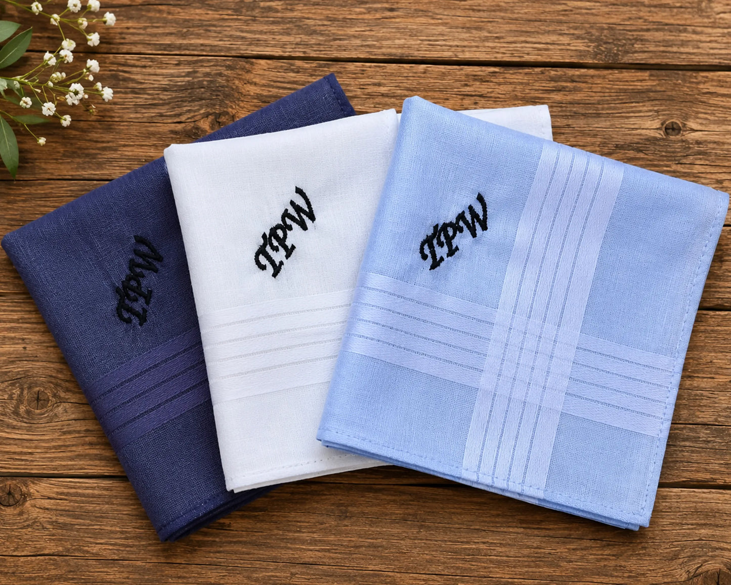 Three folded handkerchiefs in blue, white, and light blue with monograms on a wooden surface.