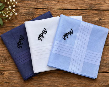 Three folded handkerchiefs in blue, white, and light blue with monograms on a wooden surface.