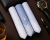 Three folded handkerchiefs with a deer emblem on a wooden surface with a watch, pen, and glass.