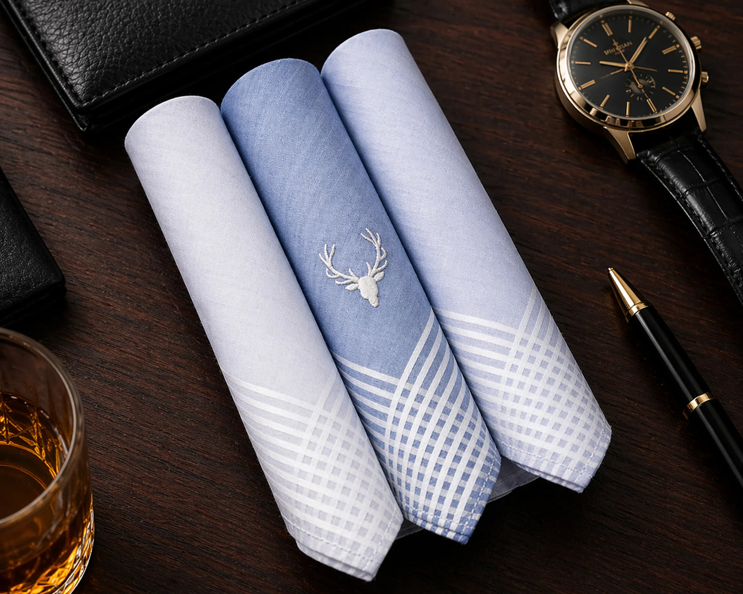 Three folded handkerchiefs with a deer emblem on a wooden surface with a watch, pen, and glass.