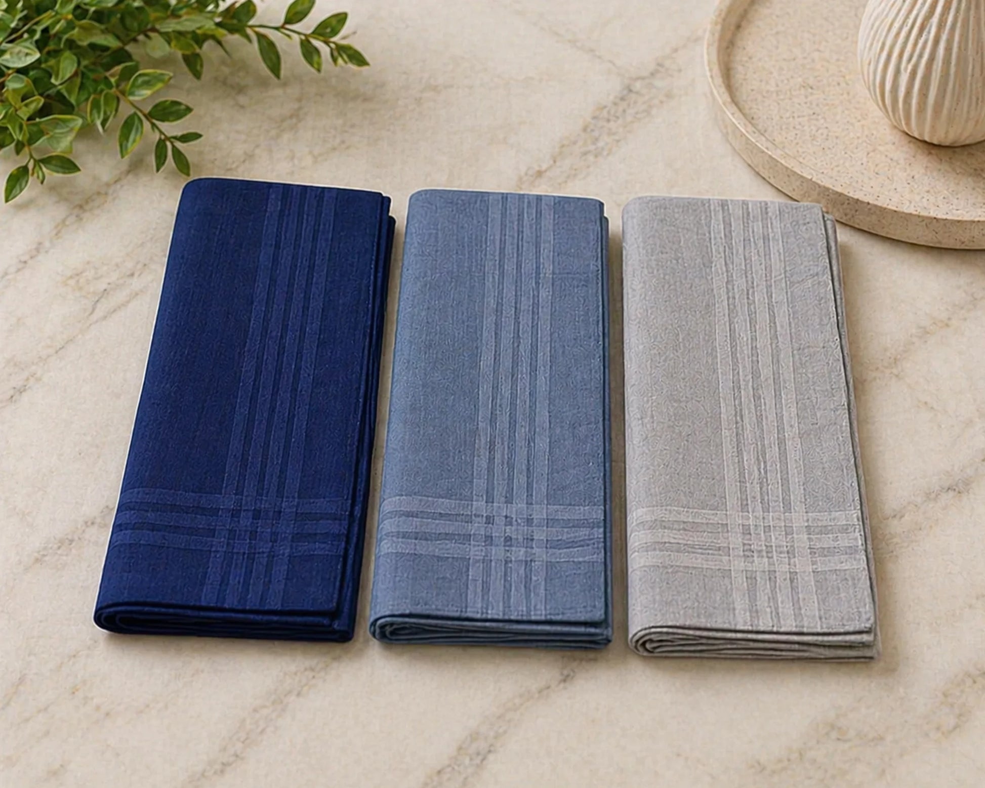 Three folded hanky in blue, gray, and green on a marble surface with a plant in the background.