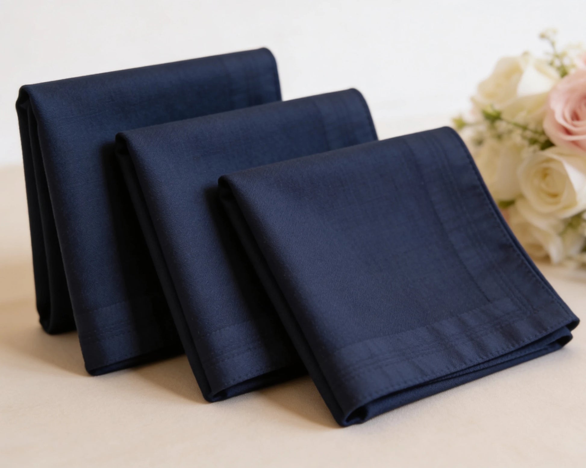 Three navy blue folded hanky on a light surface with flowers in the background
