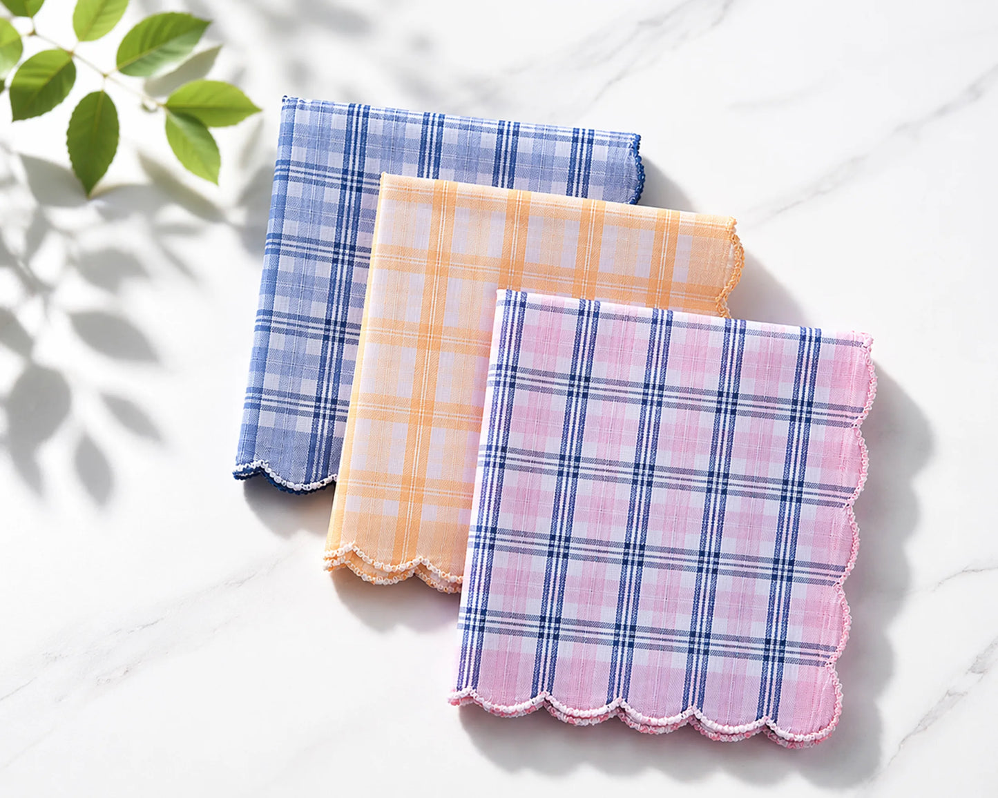 Three plaid handkerchiefs in blue, pink, and orange on a white surface with a leafy green plant in the corner.