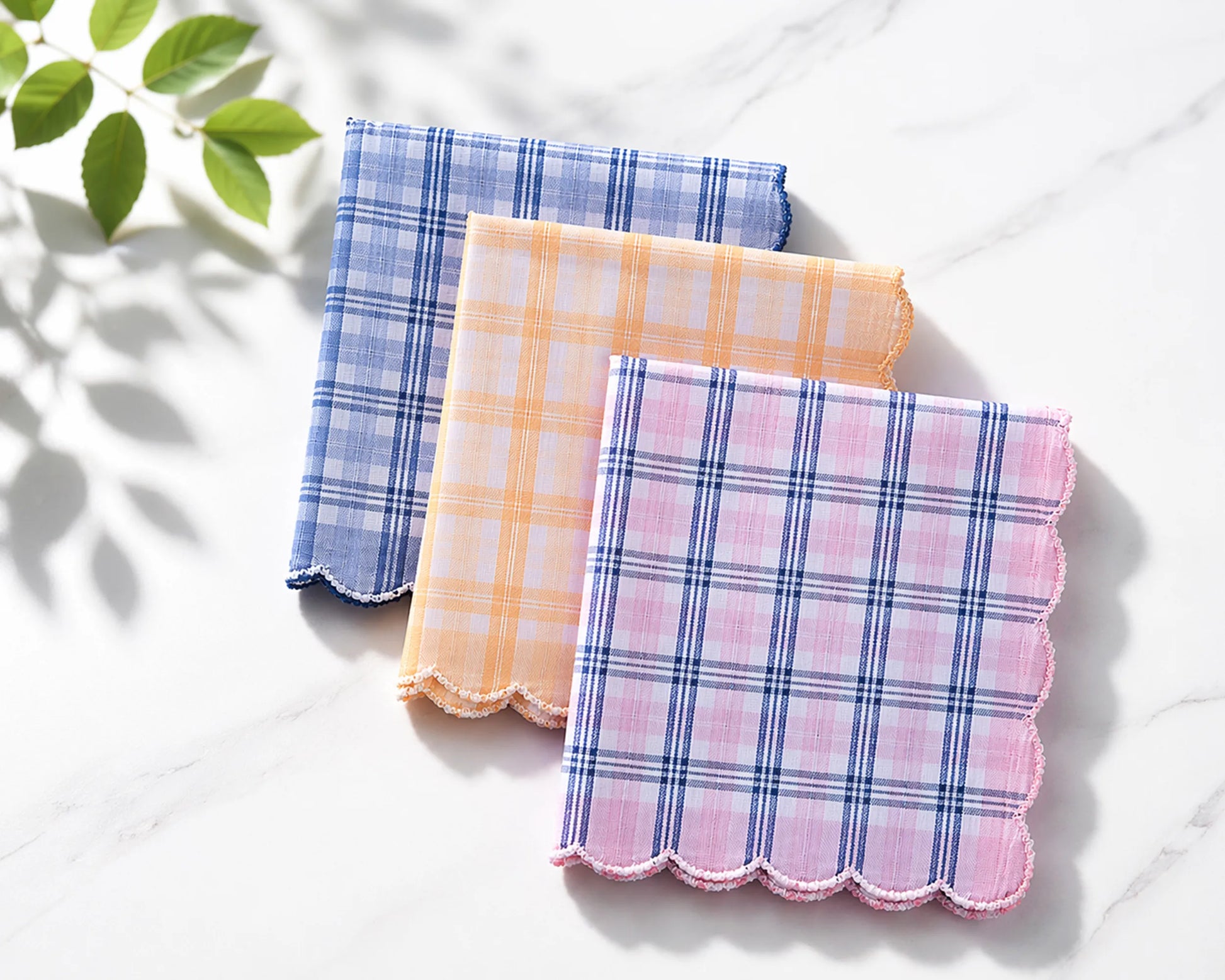 Three plaid handkerchiefs in blue, pink, and orange on a white surface with a leafy green plant in the corner.