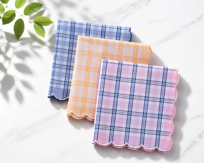 Three plaid handkerchiefs in blue, pink, and orange on a white surface with a leafy green plant in the corner.