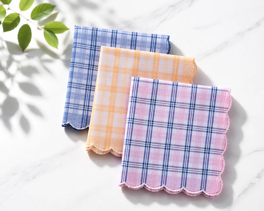 Three plaid handkerchiefs in blue, pink, and orange on a white surface with a leafy green plant in the corner.
