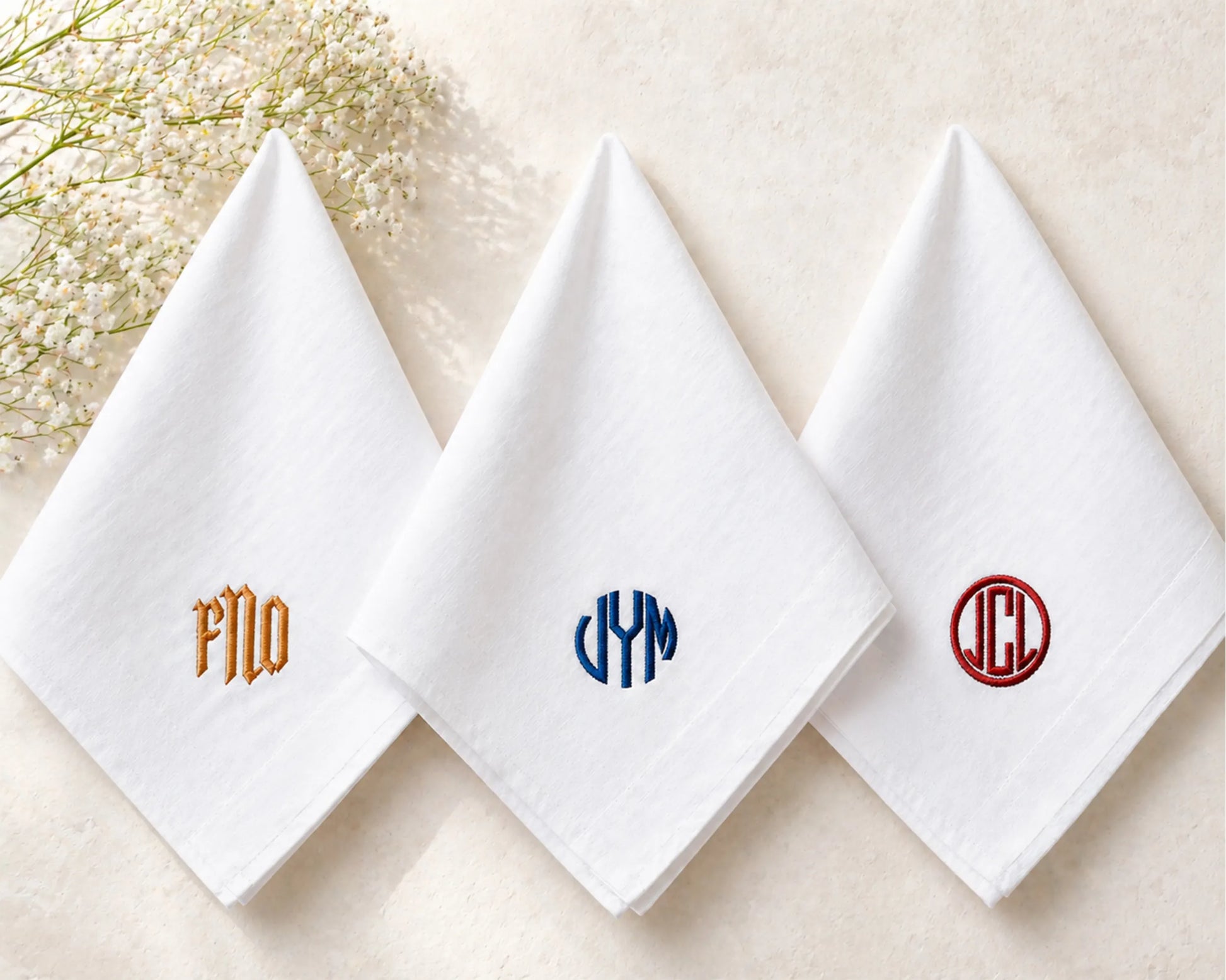 Three white handkerchiefs with embroidered monograms on a light background.
