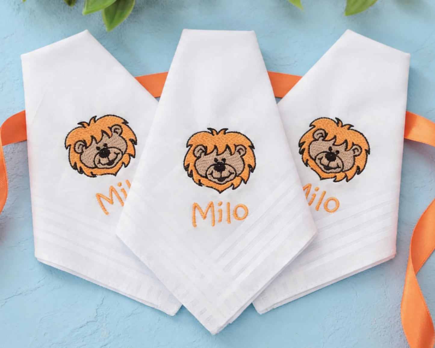 Three white handkerchiefs with lion embroidery and names embroidered on a blue background.