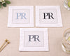 Three white napkins with 'PR' monogram on a beige surface with a glass and gold stirrer.