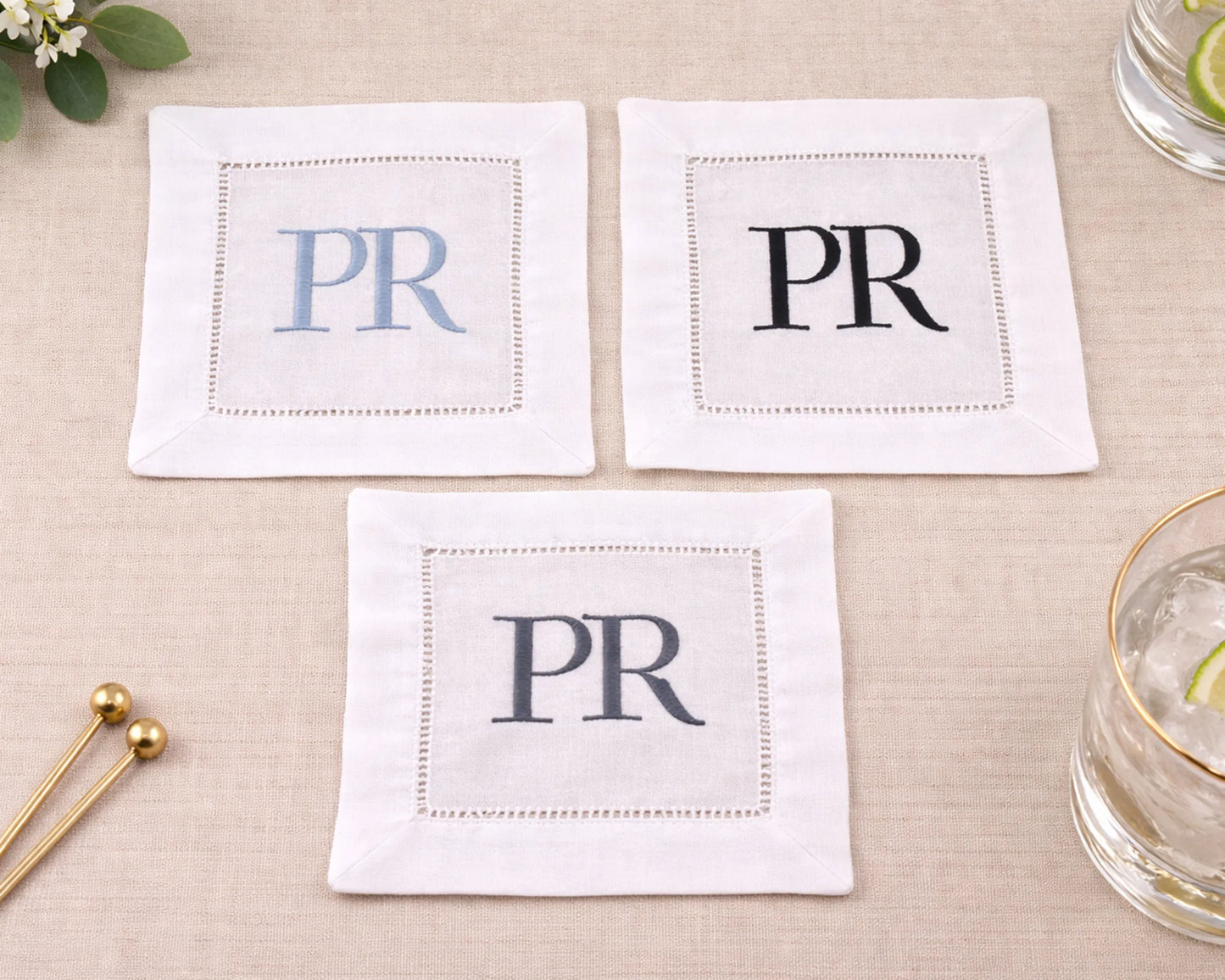 Three white napkins with 'PR' monogram on a beige surface with a glass and gold stirrer.