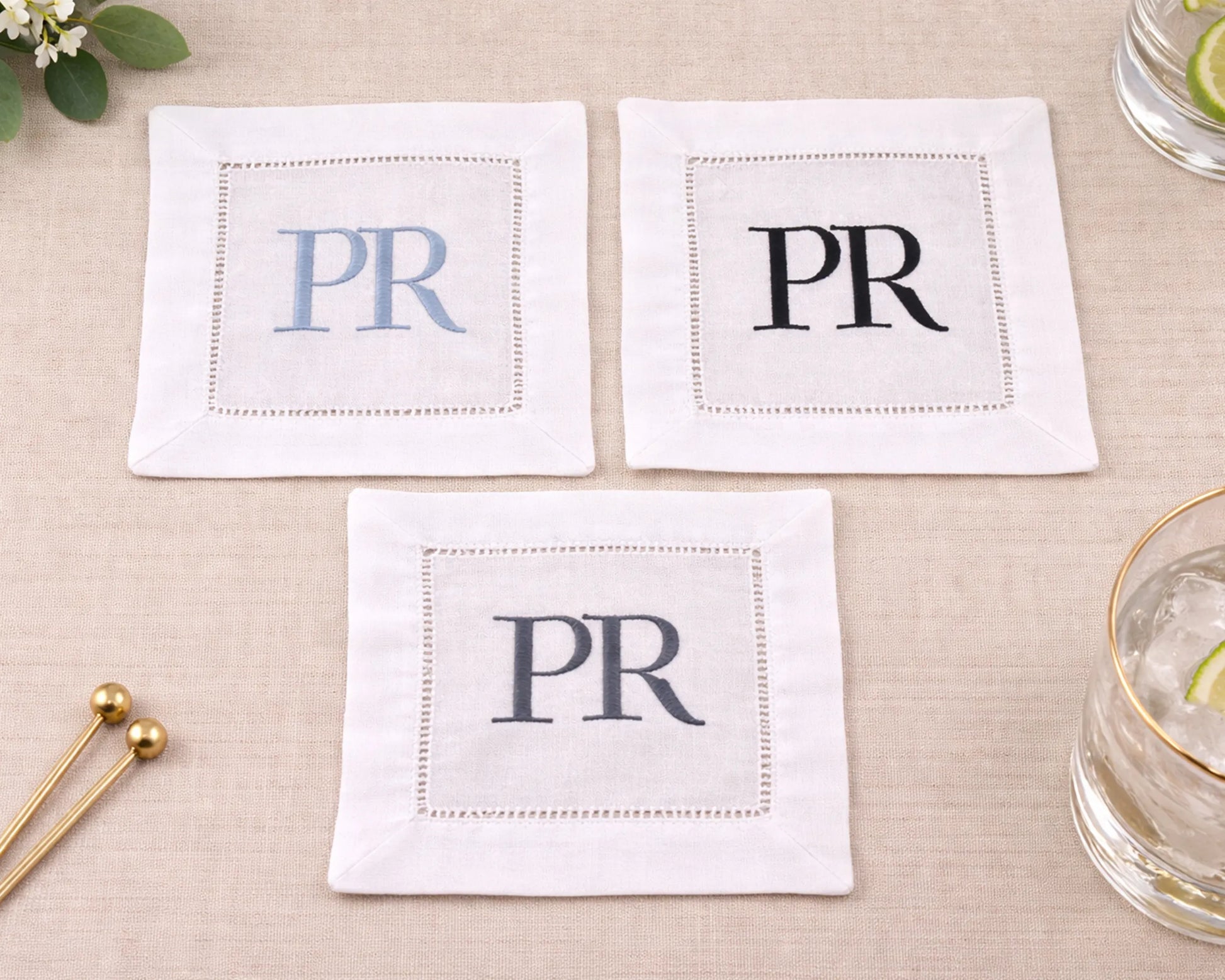 Three white napkins with 'PR' monogram on a beige surface with a glass and gold stirrer.