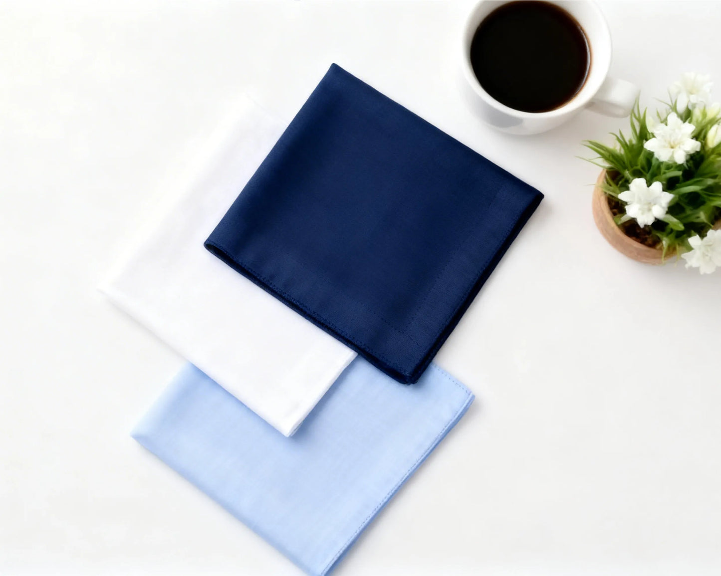 Two folded hanky, one blue and one white, on a white surface with a cup of coffee and a small plant.