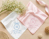 Two satin handkerchiefs with embroidered initials and dates on a wooden surface with flowers.