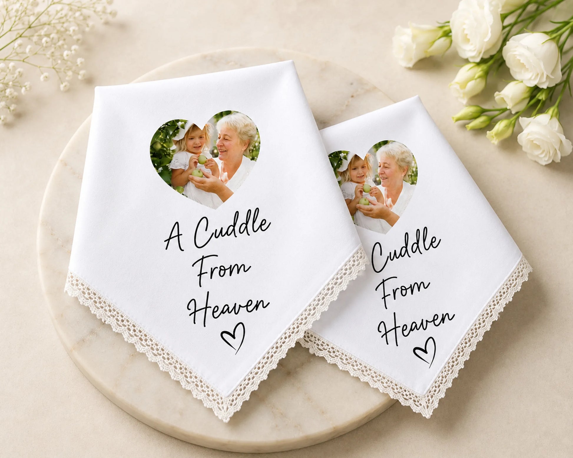 Two white handkerchiefs with lace edges featuring heart-shaped photos and text 'A Cuddle From Heaven' on a beige surface with flowers.