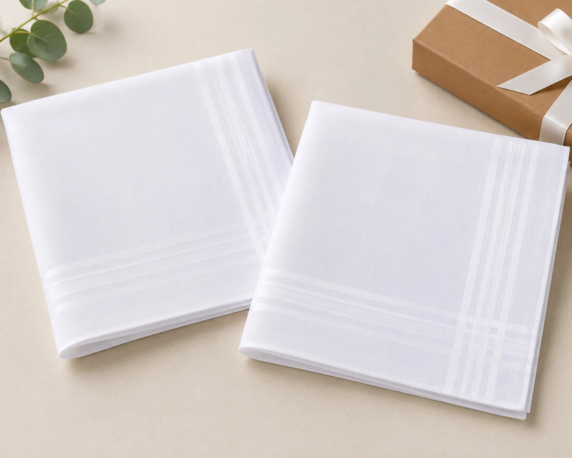Two white hanky on a beige surface with a gift box and greenery in the background.