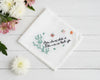White cotton handkerchief featuring centered green foliage embroidery and black calligraphic text on a neutral background