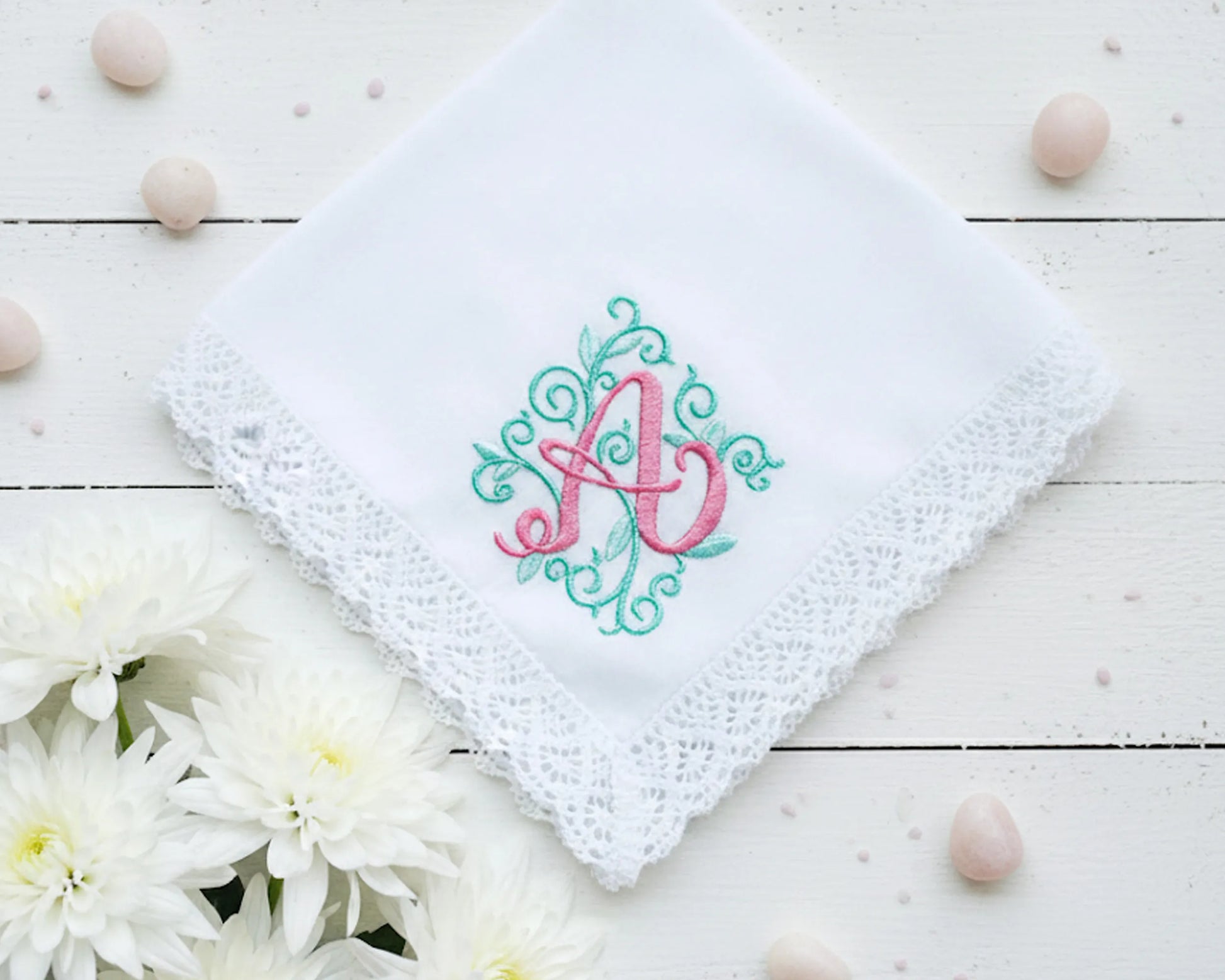 A white lace-edged handkerchief featuring a decorative pink A initial with teal vine embroidery on a white wooden surface