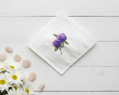 Detailed violet hydrangea embroidery with green leaves on a white cotton handkerchief displayed with smooth stones