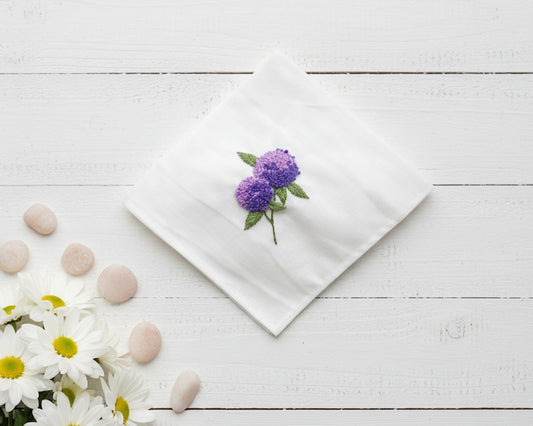 Detailed violet hydrangea embroidery with green leaves on a white cotton handkerchief displayed with smooth stones