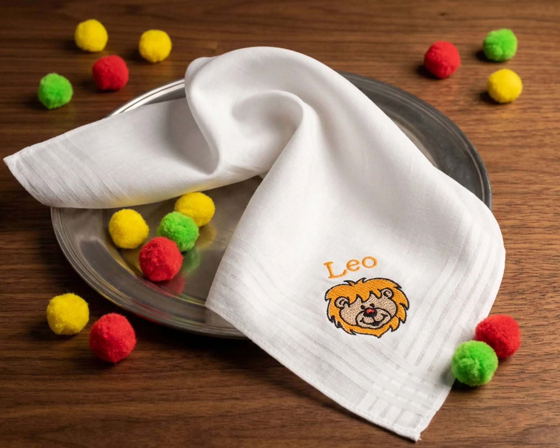 White baby blanket with embroidered lion design and 'Leo' text, placed on a wooden surface with colorful pom-poms.