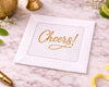 White cocktail napkin with 'Cheers!' in gold on a marble surface with decorative items.