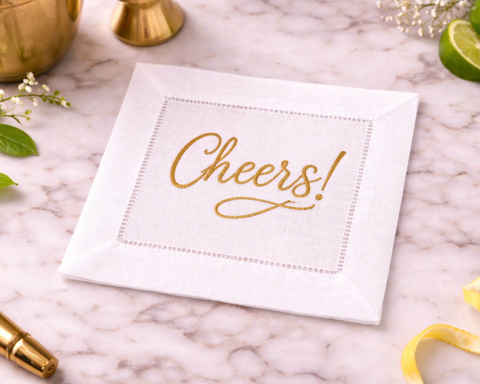 White cocktail napkin with 'Cheers!' in gold on a marble surface with decorative items.